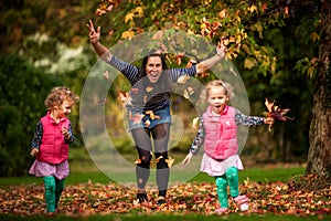 Mother and identical twins having fun with autumn leaves under the tree in the park, blond cute curly girls, happy family