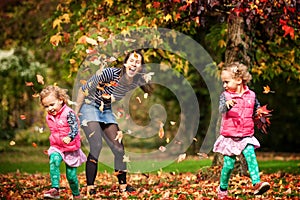 Mother and identical twins having fun with autumn leaves under the tree in the park, blond cute curly girls, happy family