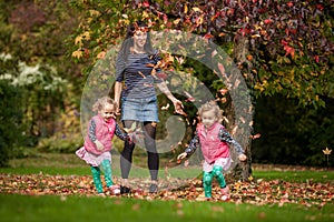 Mother and identical twins having fun with autumn leaves under the tree in the park, blond cute curly girls, happy family