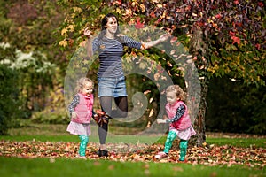 Mother and identical twins having fun with autumn leaves under the tree in the park, blond cute curly girls, happy family
