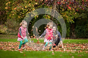 Mother and identical twins having fun with autumn leaves under the tree in the park, blond cute curly girls, happy family