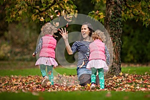 Mother and identical twins having fun with autumn leaves under the tree in the park, blond cute curly girls, happy family