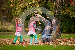 Mother and identical twins having fun with autumn leaves under the tree in the park, blond cute curly girls, happy family