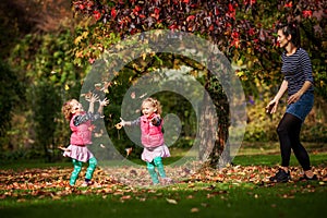 Mother and identical twins having fun under the tree with autumn leaves in the park, blond cute curly girls, happy family