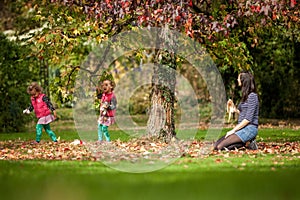 Mother and identical twins having fun under the tree with autumn leaves in the park, blond cute curly girls, happy family