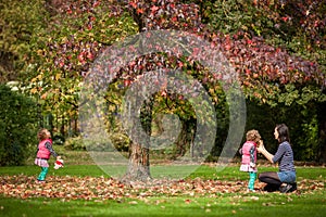 Mother and identical twins having fun under the tree with autumn leaves in the park, blond cute curly girls, happy family