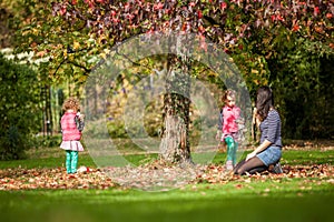 Mother and identical twins having fun under the tree with autumn leaves in the park, blond cute curly girls, happy family