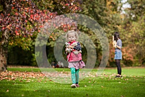 Mother and igirl having fun in the park, blond cute curly girls walking away from mother with her toys, happy family