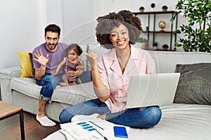 Mother of interracial family working using computer laptop at home pointing to the back behind with hand and thumbs up, smiling