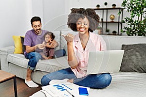Mother of interracial family working using computer laptop at home smiling with happy face looking and pointing to the side with