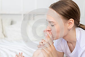 Mother kisses baby`s feet and sniffs them on the bed at home, happy motherhood