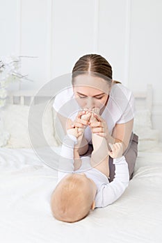 Mother kisses baby`s feet and sniffs them on the bed at home, happy motherhood