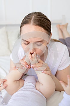 Mother kisses baby`s feet and sniffs them on the bed at home, happy motherhood