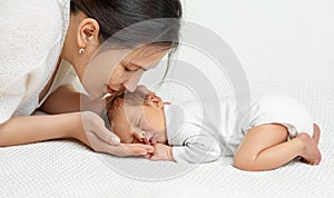 Mother kissing Baby sleeping on white Blanket. Smiling Mom holding Newborn Head in Hands. Infant Side view lying down on Stomach.