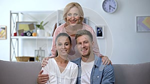 Mother-in-law hugging young couple showing hands with engagement rings at home