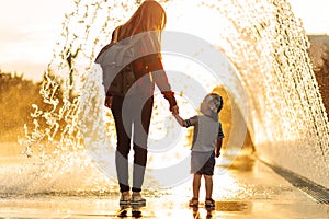 Mother and little boy are walking in the park, mom and son are walking holding hands near the fountain at sunset