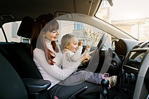 Mother with little child girl on her knees shows how to drive a car and what is steering wheel. Mom and baby having fun