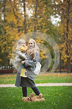 Mother and little daughter blonde with Teddy bear standing on green grass in autumn park