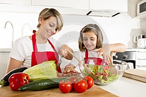 Mother and little daughter cooking together with cook apron preparing salad at home kitchen