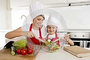 Mother and little daughter cooking together with hat apron preparing salad at home kitchen