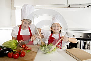 Mother and little daughter cooking together with hat apron preparing salad at home kitchen