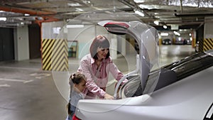 Mother and little daughter putting colorful bags with clothes into trunk of car.