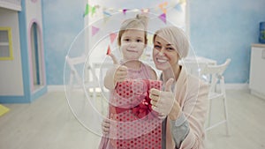 Mother and little daughter sitting in front of the camera with birthday present, smiling and showing thumbs