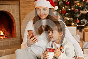 Mother and little daughter with smart phone posing near fireplace, sitting on floor and look at screen of mobile phone, child with
