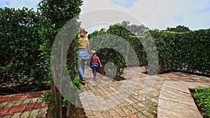 Mother Little Girl Walk on Flowerbed with Angel Statue in Park