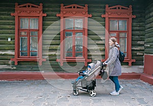 Mother and the little son in a stroller walk on the city street along the big old wooden house