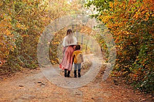 Mother and little son on walk in autumn forest, walking along path covered with yellow tree leaves
