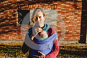 Mother lovingly carrying her newborn baby in a sling scarf during a sunset stroll in front of red brick building