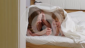 Mother lying on bed under duvet blanket with daughter kid telling secrets to each other, smilling