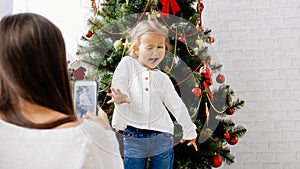 Mother making photo with smart phone of her little daughter near christmas tree