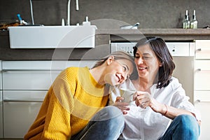 Mother and mature daughter in kitchen, sitting on floor and talking. Closeness of mom and her adult offspring. photo