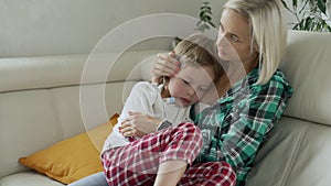 Mother measuring temperature of her ill kid at background. Sick child with high fever lying at sofa at home