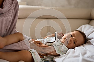 Mother measuring temperature of her ill kid at background. Sick child with high fever lying at sofa at home.