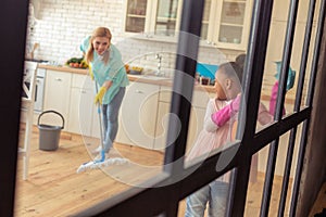 Mother mopping the floor and looking at her helpful daughter