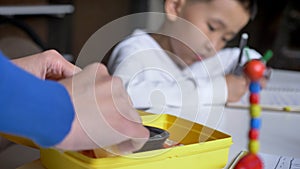 Mother packing lunch for kid while little asian boy doing homework and sitting behind table, book shelves and brick wall