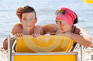 Mother pat her teenage son on the back while lying on yellow sun lounger on coastline, touching hair and looking at boy