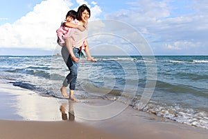 Mother piggybacking her kid on beach, playing together on summer holiday vacation, joyful parent with child boy spending time on