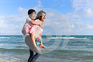 Mother piggybacking her kid on beach, playing together on summer holiday vacation, joyful parent with child boy spending time on