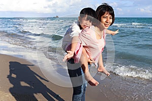 Mother piggybacking her kid on beach, playing together on summer holiday vacation, joyful parent with child boy spending time on