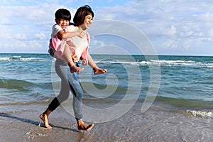 Mother piggybacking her kid on beach, playing together on summer holiday vacation, joyful parent with child boy spending time on