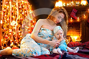 Mother playing with baby while sitting near Christmas tree and fireplace in decorated living room