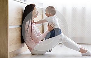 Mother playing with cute baby, sitting on floor in kitchen