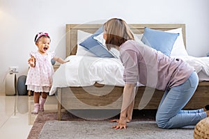 Mother is playing peek a boo or hide and seek with her little baby toddler in the bedroom while the girl is laughing in happiness