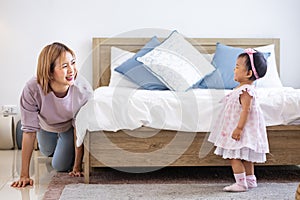 Mother is playing peek a boo or hide and seek with her little baby toddler in the bedroom while the girl is laughing in happiness