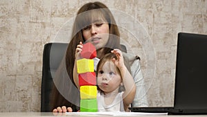 Mother plays with her child in multi-colored cubes at work in the office. Busy woman working on laptop with baby on