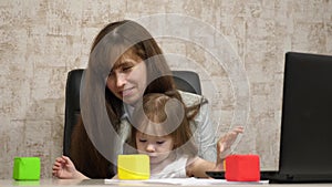 Mother plays with her child in multi-colored cubes at work in the office. Busy woman working on laptop with baby on